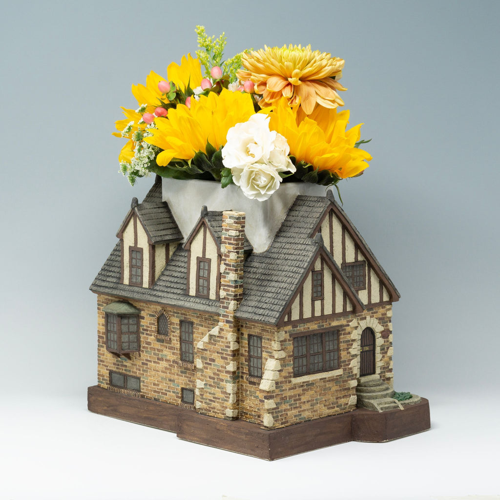 A handcrafted and highly detailed vase in the shape of a home containing a bouquet of yellow and white flowers.